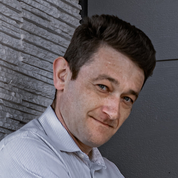 ciemnowłosy mężczyzna w średnim wieku w jasnej koszuli. Opiera się o ścianę budynku.